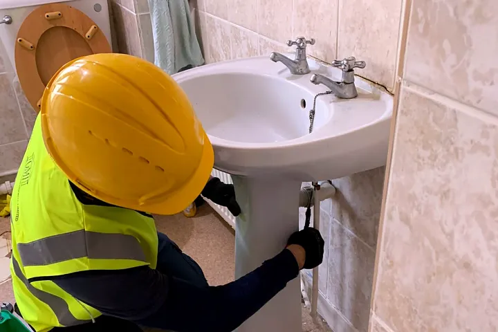 An engineer fitting the waste and tap connections under a pedestal basin in a tiled bathroom